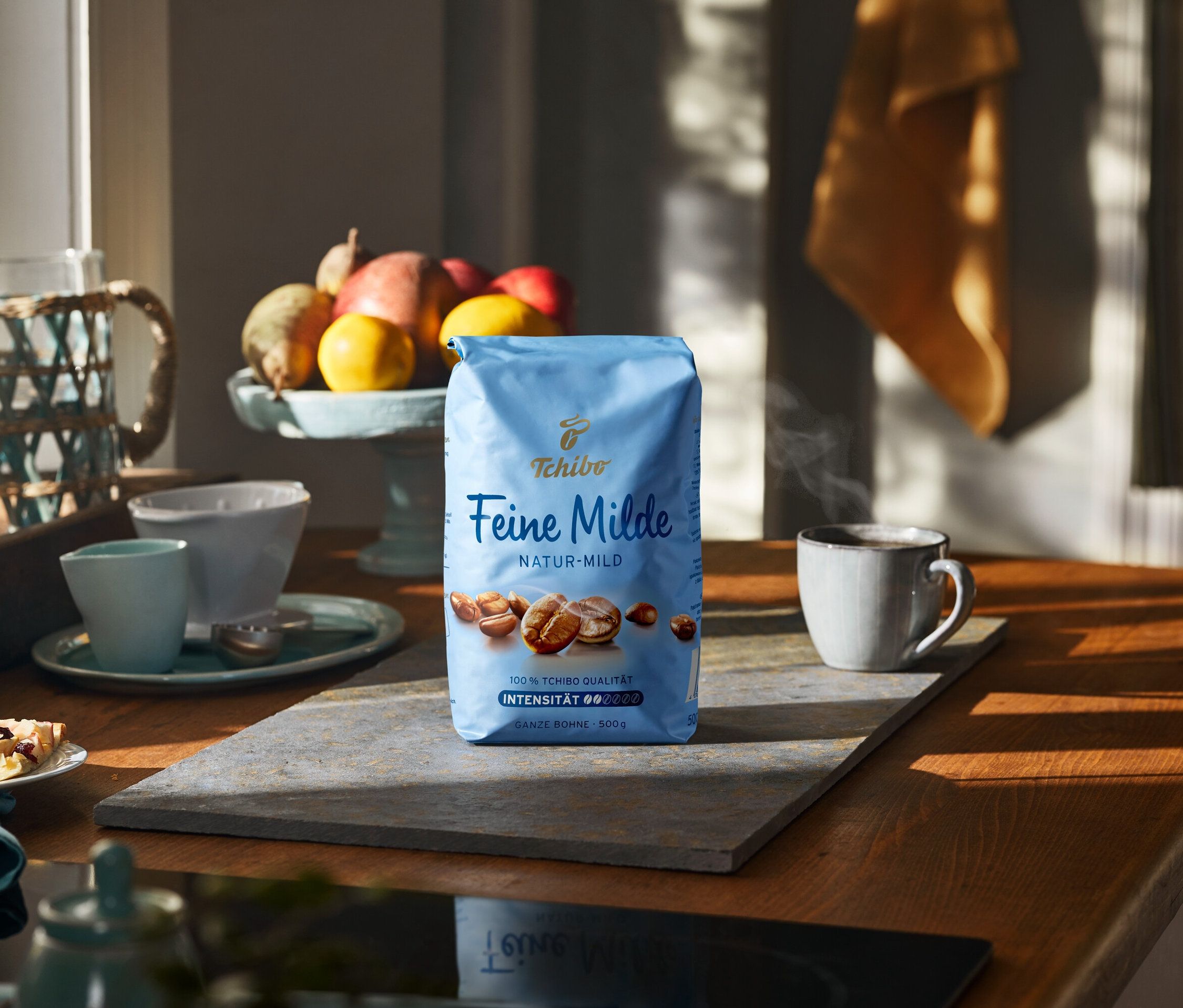 Eine 500-g-Packung Feine Milde Kaffee auf einer Steinplatte neben einer Tasse und Früchten.