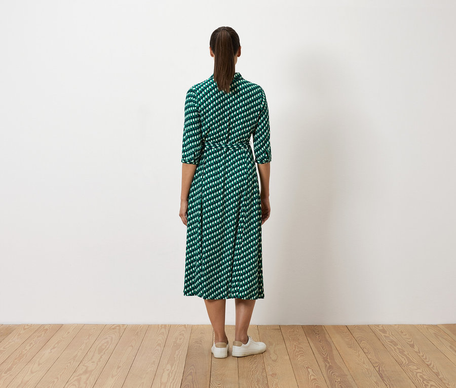 Eine Frau steht mit dem Rücken zur Kamera und trägt ein grünes Kleid mit weißem Muster und weiße Turnschuhe.
