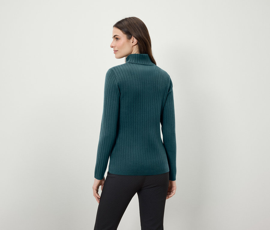 Frau mit dem Rücken zur Kamera trägt einen grünen Rollkragenpullover und eine schwarze Hose.