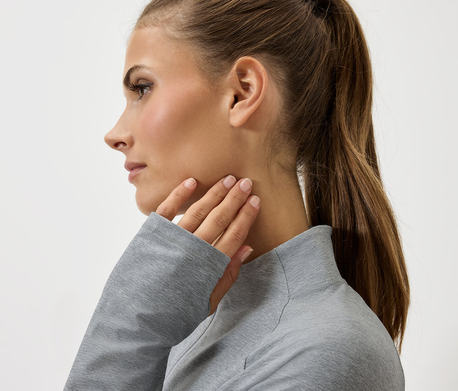 Profil einer Frau mit braunen Haaren im Pferdeschwanz, die ihren Hals mit einer Hand in einem grauen Pullover berührt.