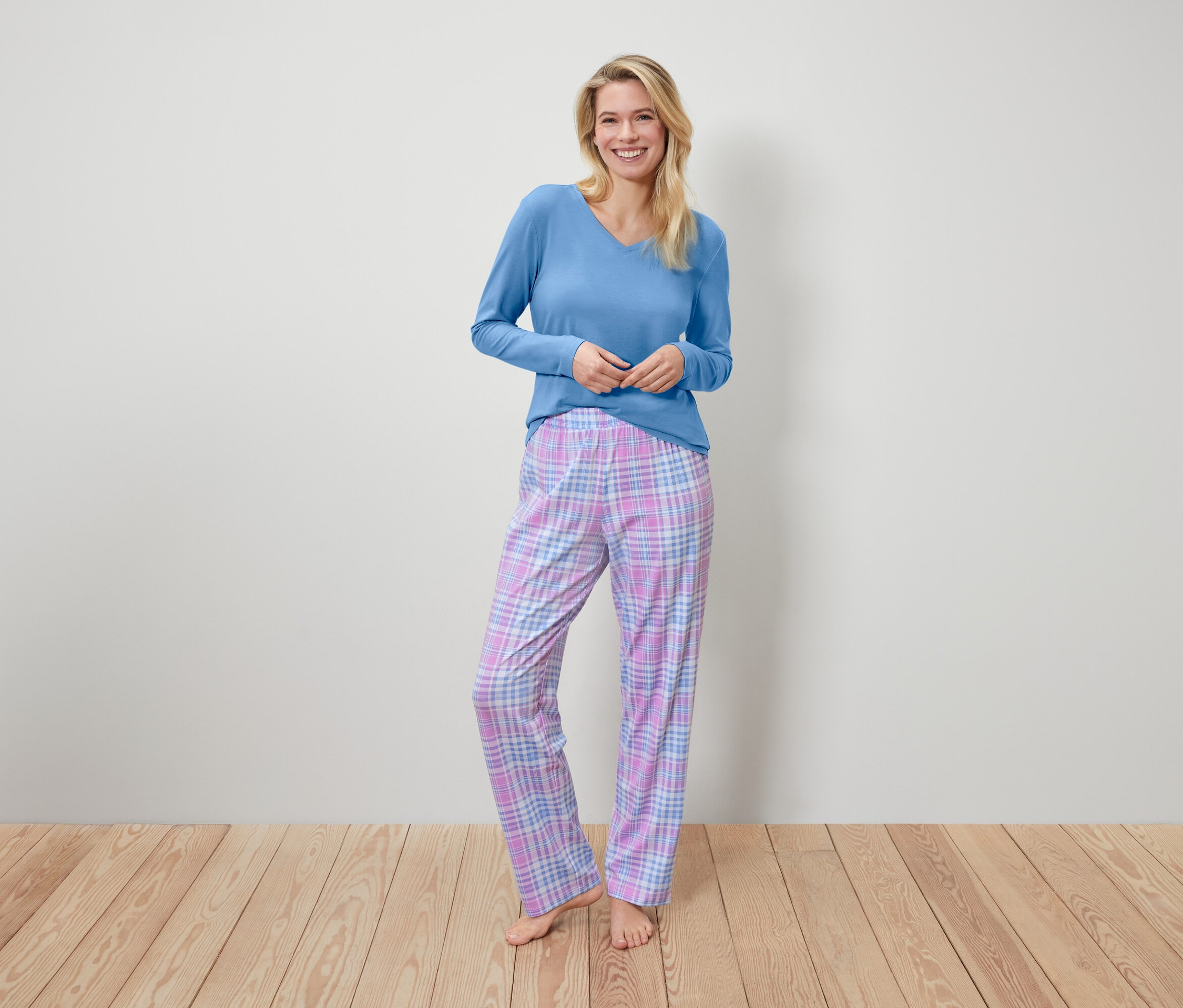 Eine lächelnde Frau steht und trägt ein blaues Langarmshirt und eine karierte Pyjamahose.