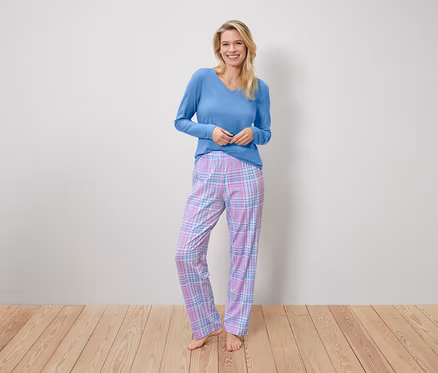 Eine lächelnde Frau steht und trägt ein blaues Langarmshirt und eine karierte Pyjamahose.