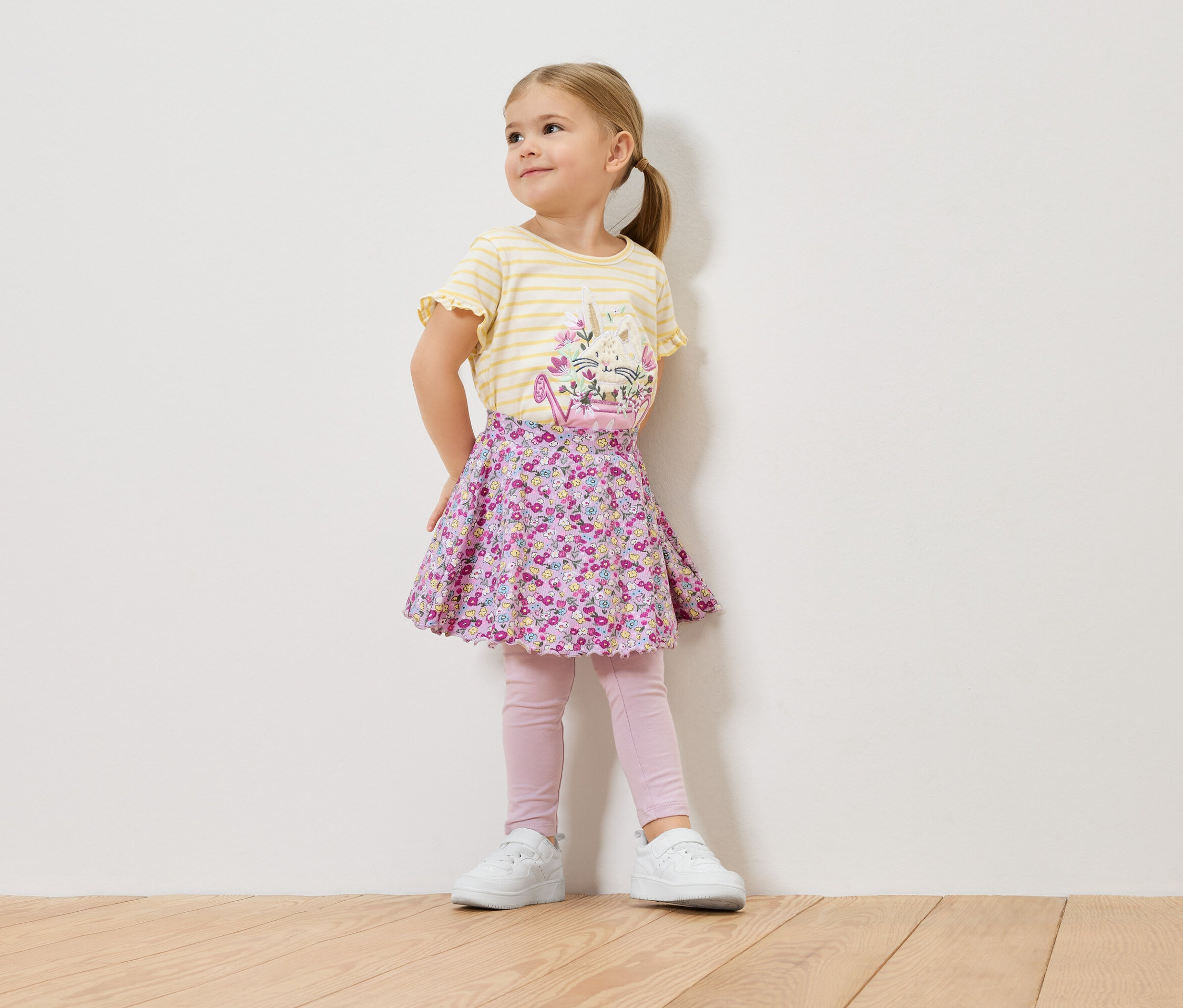 Ein kleines Mädchen mit blonden Haaren trägt ein gelb-gestreiftes Shirt mit einem Blumenmuster, einen geblümten Rock, rosa Leggings und weiße Turnschuhe.