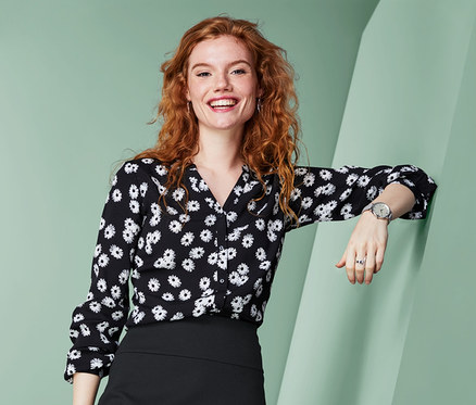 Eine Frau lächelt, an eine Wand gelehnt, mit einer schwarzen Bluse mit Blumenmuster und einem schwarzen Jerseyrock.