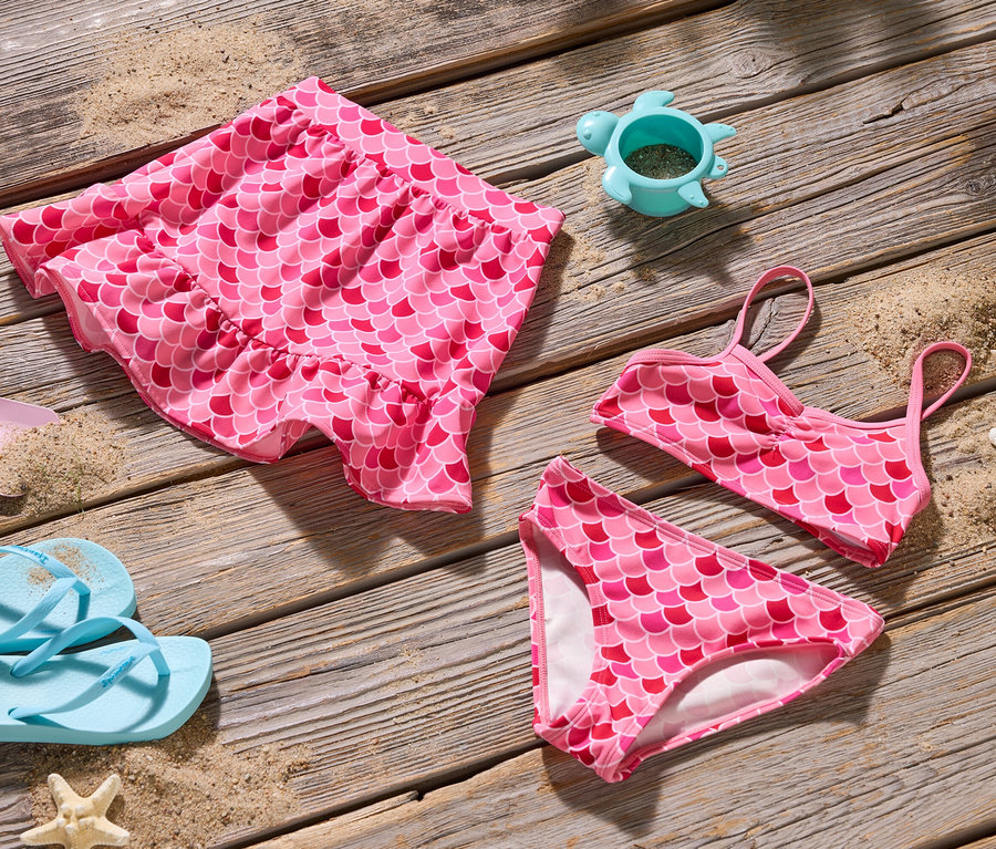 Draufsicht auf ein rosafarbenes Meerjungfrauen-Bikini-Set, einen Rock, eine Schaufel, Flip-Flops und eine Schildkröten-Sandform auf Holz.
