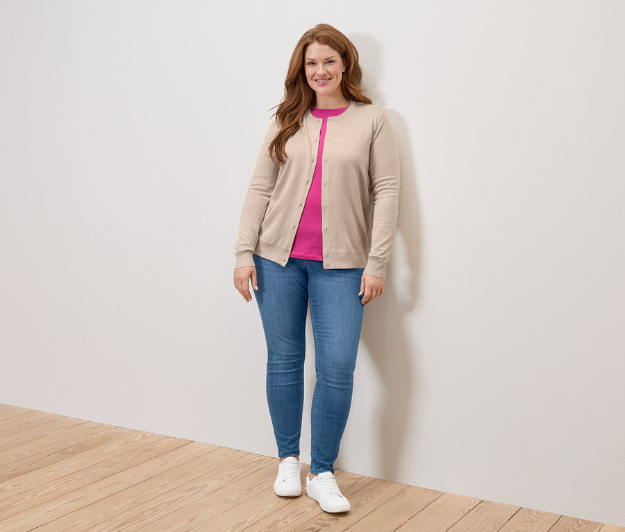 Eine Frau mit langen, roten Haaren steht vor einer Wand und trägt eine beige Strickjacke, ein pinkfarbenes T-Shirt, Jeans und weiße Turnschuhe.