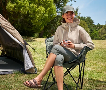 Frau sitzt im Tambu »ASANA« Camping-Faltstuhl, hält eine Tasse und trägt 3/4-Outdoor-Leggings und einen Kapuzenpullover.