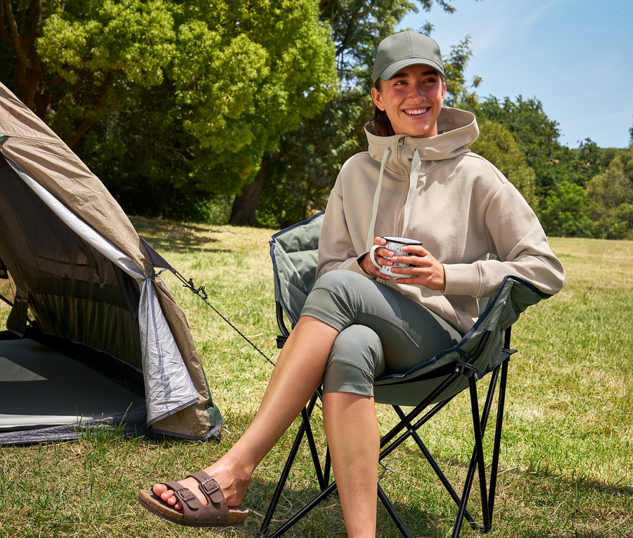 Frau sitzt im Tambu »ASANA« Camping-Faltstuhl, hält eine Tasse und trägt 3/4-Outdoor-Leggings und einen Kapuzenpullover.