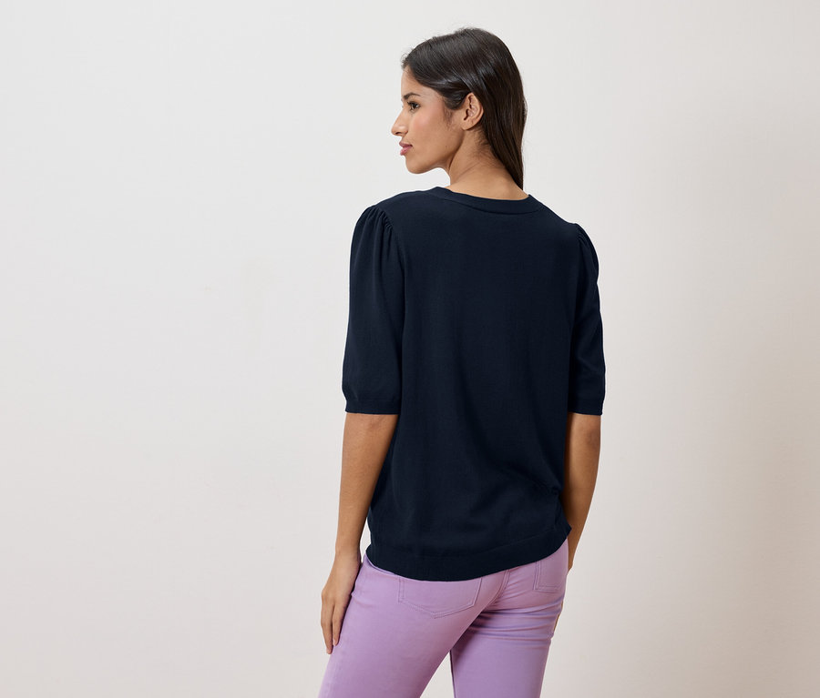 Rückansicht einer Frau mit dunklen Haaren, die einen marineblauen Pullover und lila Hosen trägt.