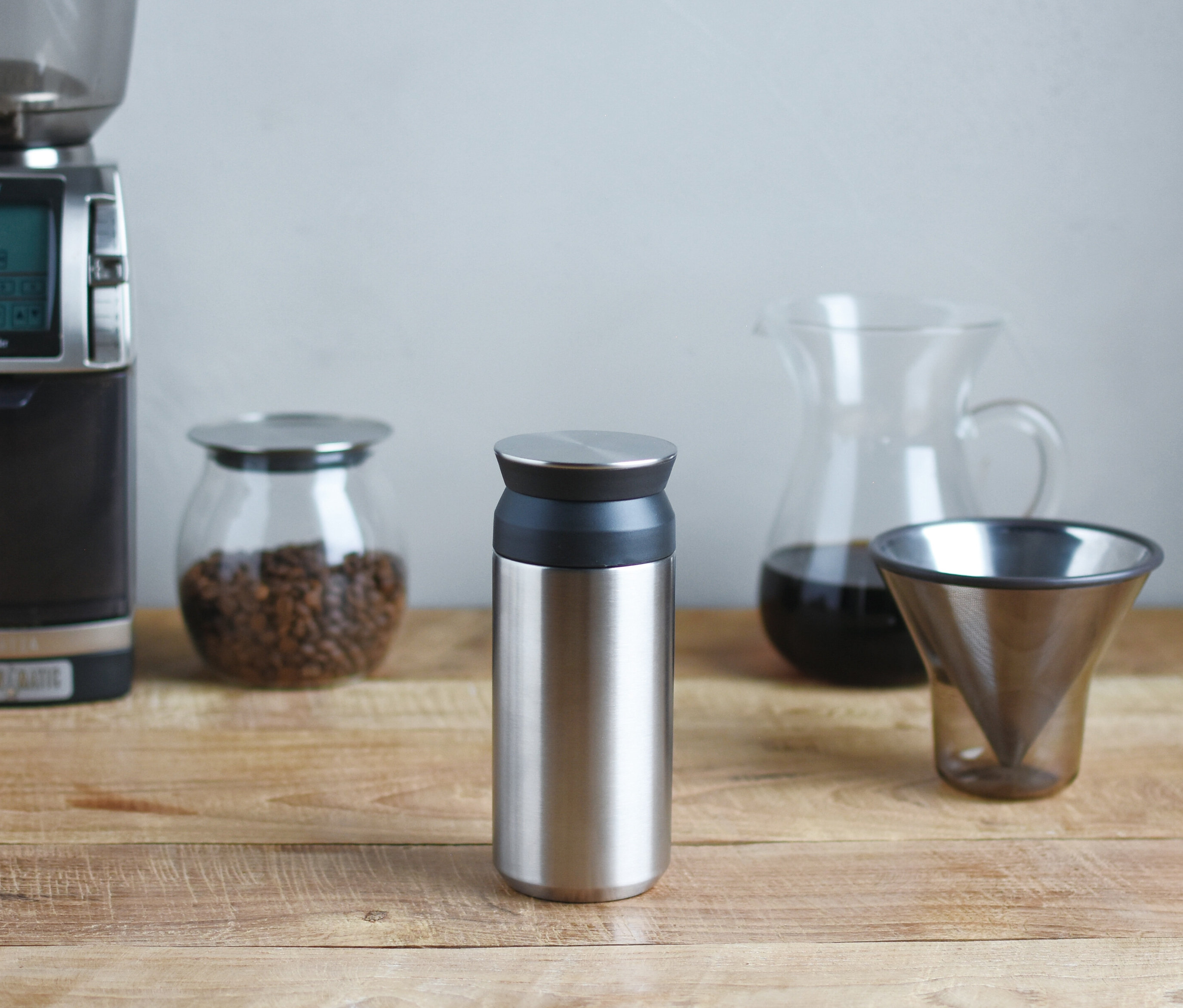 Ein silberner Thermobecher steht auf einem Holztisch, umgeben von einer Kaffeemühle, Kaffeebohnen, einer Karaffe und einem Kaffeefilter.