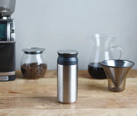 Ein silberner Thermobecher steht auf einem Holztisch, umgeben von einer Kaffeemühle, Kaffeebohnen, einer Karaffe und einem Kaffeefilter.