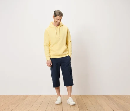Mann mit gelbem Kapuzenpullover und blauen Cargo-Shorts, der auf einem Holzboden steht.