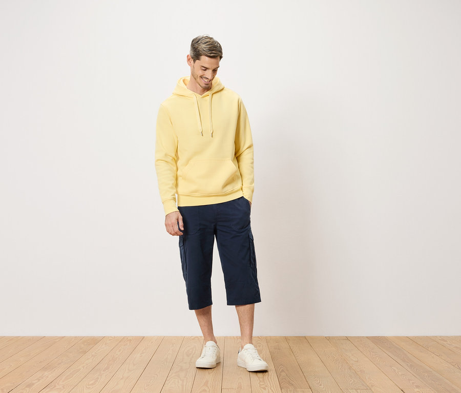 Mann mit gelbem Kapuzenpullover und blauen Cargo-Shorts, der auf einem Holzboden steht.