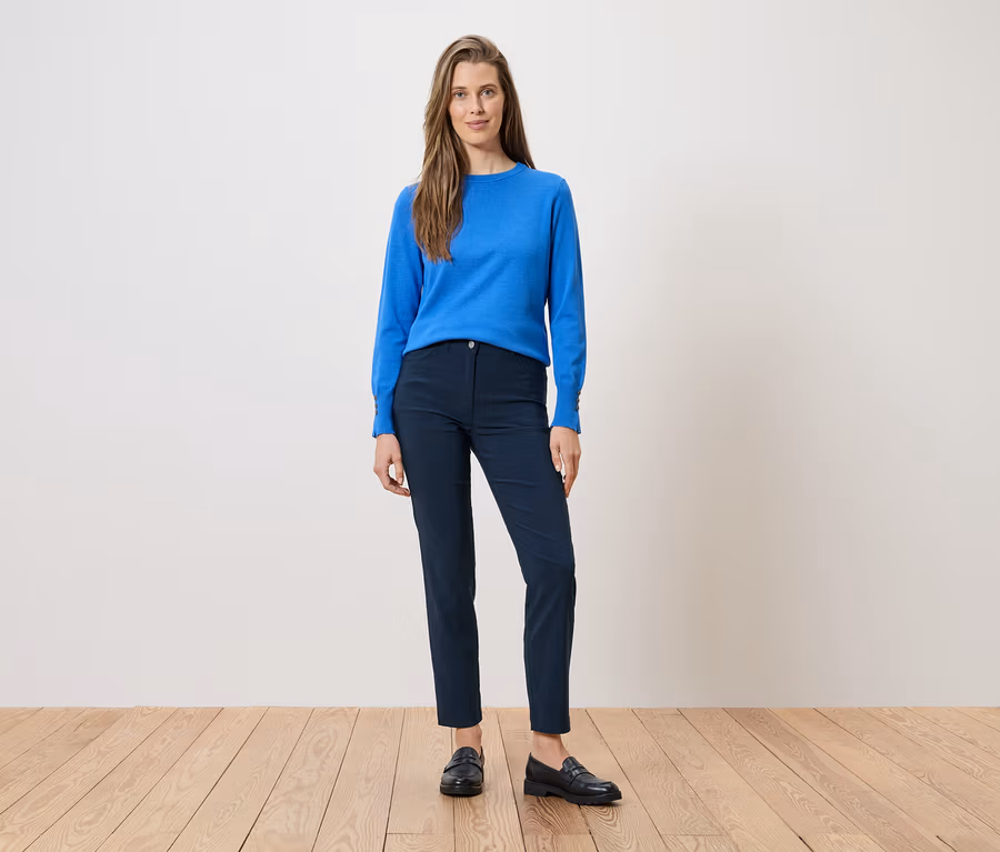 Frau in blauer Bluse und marineblauer Hose, dazu schwarze Slipper. Sie steht auf einem Holzboden vor einer weißen Wand.