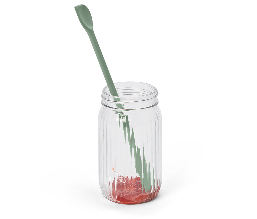 Ein grüner Löffel steckt in einem gerippten Glas mit etwas roter Marmelade am Boden.