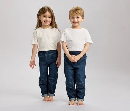 Zwei Kinder mit blonden und braunen Haaren stehen barfuß nebeneinander und tragen weiße T-Shirts und blaue Jeans mit Bärenmotiven.