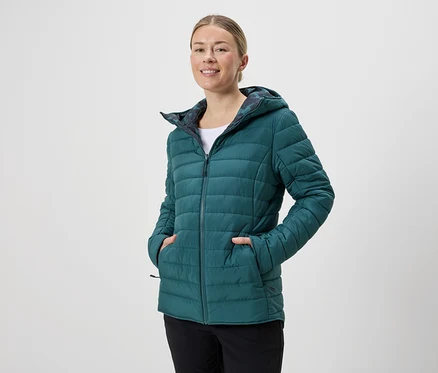 Frau mit Kapuze trägt eine smaragdgrüne Steppjacke mit Reißverschluss und schwarzen Hosen vor einem neutralen Hintergrund.