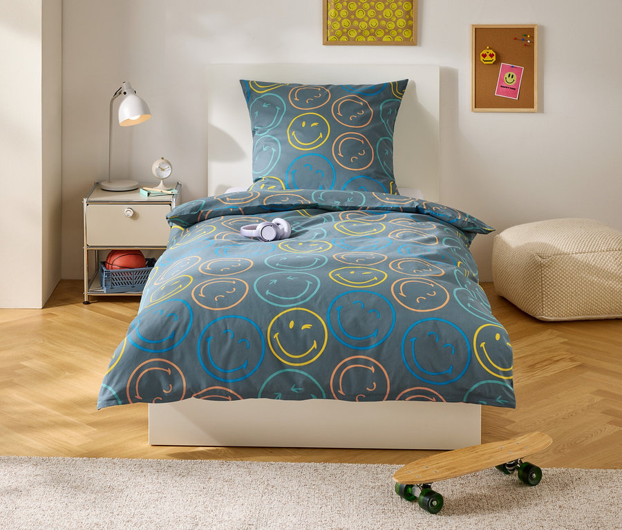 Einzelbett mit dunkelgrauer Bettwäsche, bunten Smiley-Gesichtern. Nachttisch mit Lampe, Korb mit Basketball. Skateboard auf Teppich vor dem Bett.