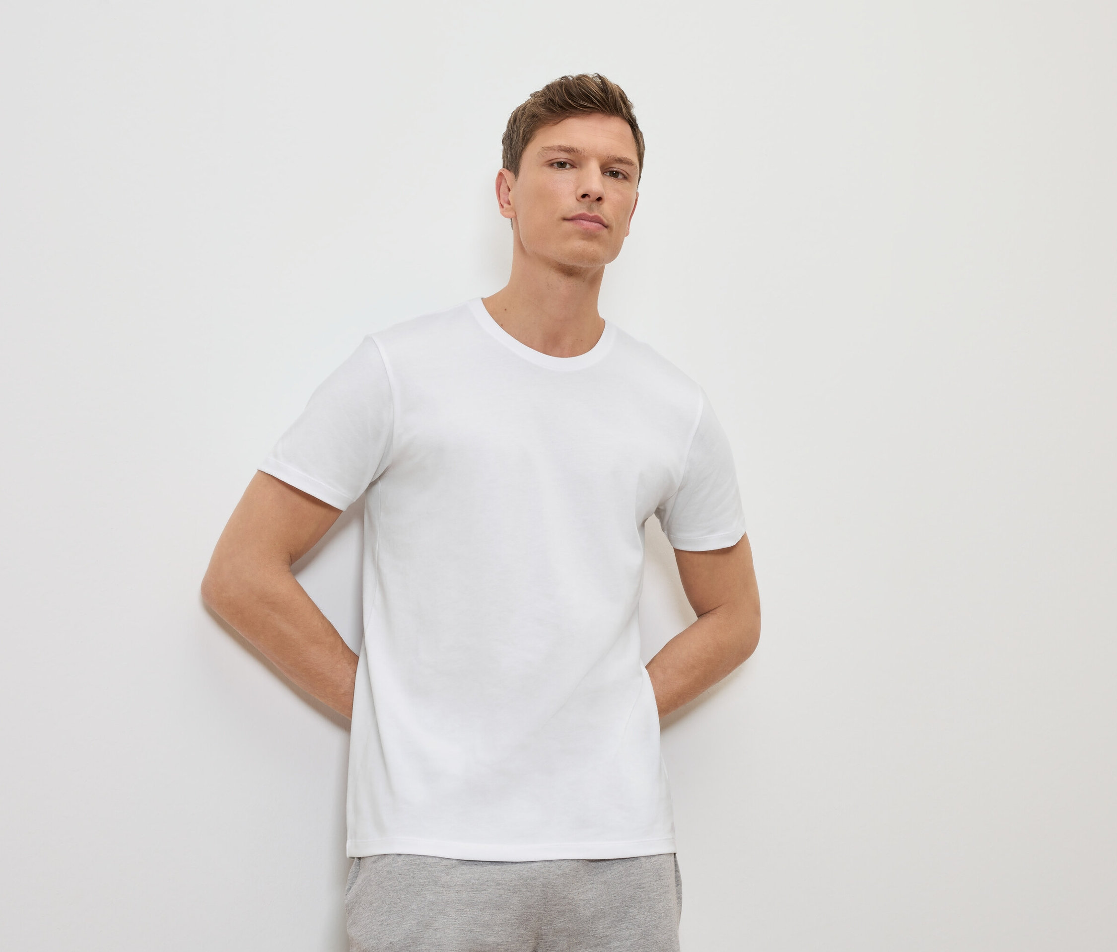 Mann lehnt an einer weißen Wand und trägt ein weißes T-Shirt und graue Jogginghose.