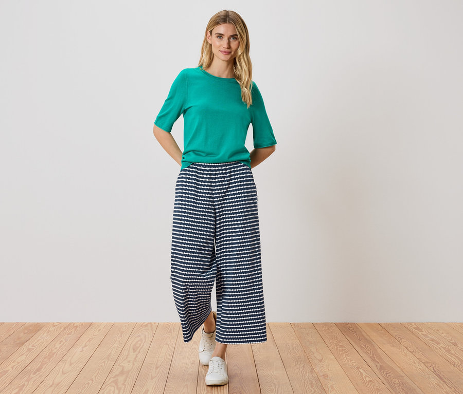Frau steht mit Händen in den Hosentaschen, trägt ein grünes T-Shirt, eine blau-weiß gestreifte Hose und weiße Schuhe.