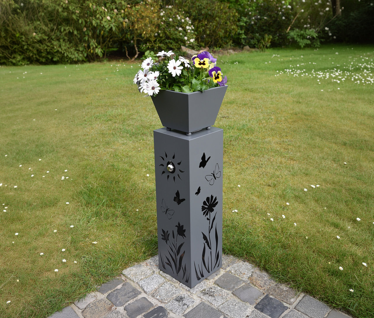 Graue Pflanzsäule mit Aussparungen von Schmetterlingen, Blumen und Sonne, darauf ein Topf mit weißen und lila Stiefmütterchen.