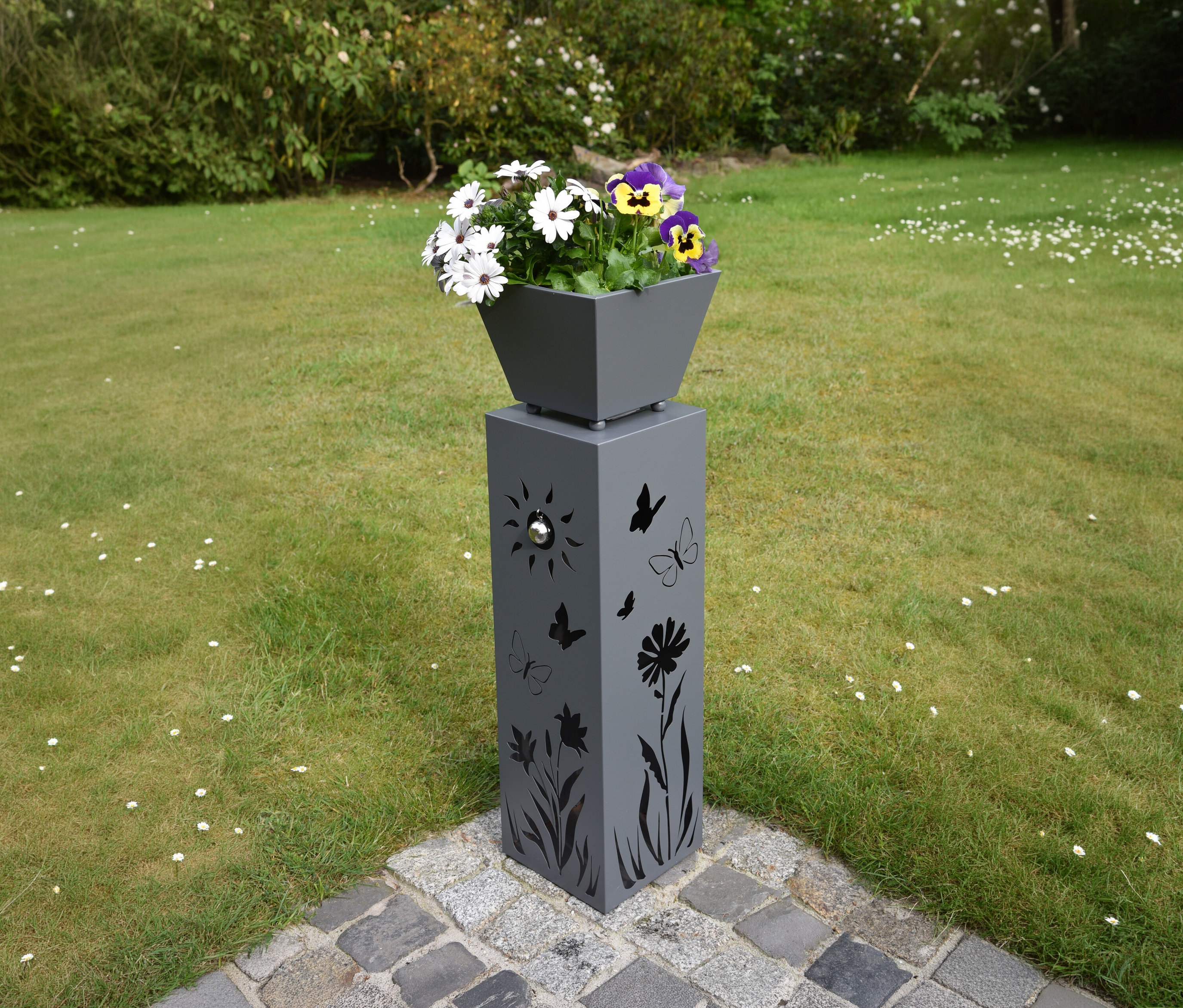 Graue Pflanzsäule mit Aussparungen von Schmetterlingen, Blumen und Sonne, darauf ein Topf mit weißen und lila Stiefmütterchen.