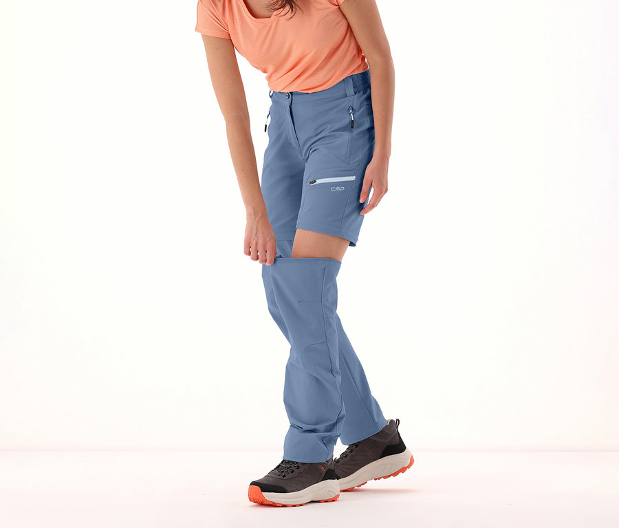 Frau befestigt Hosenbein an CMP Damen-Zip-off-Hose in Blau. Sie trägt ein orangefarbenes T-Shirt und grau-schwarze Schuhe.