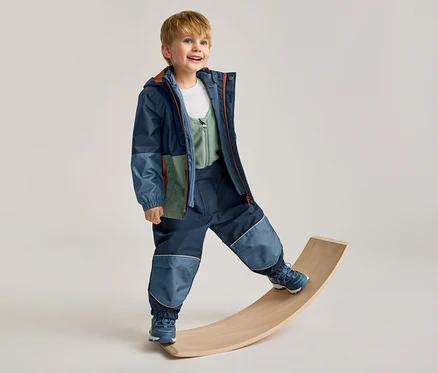 Lächelnder Junge in einer mehrfarbigen Jacke und dunkelblauen Hose mit hellblauen Patches, der auf einem Holzbrett balanciert.