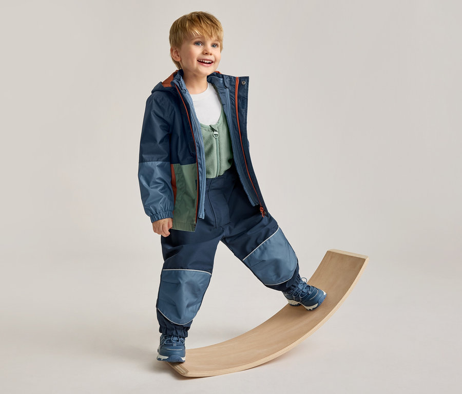 Lächelnder Junge in einer mehrfarbigen Jacke und dunkelblauen Hose mit hellblauen Patches, der auf einem Holzbrett balanciert.