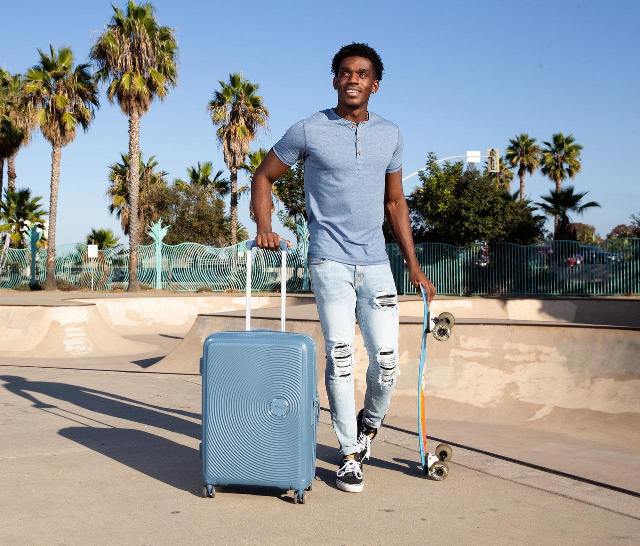 Mann mit American Tourister »Soundbox« Hartschalen-Koffer in Stone Blue und Skateboard in Skatepark.