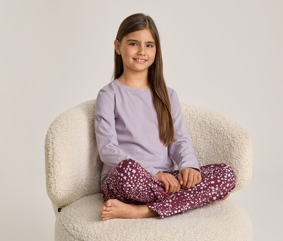 Ein lächelndes Mädchen sitzt im Schneidersitz auf einem cremefarbenen Sessel. Sie trägt ein hellviolettes Langarmshirt und dunkelrote Hosen mit weißen Sternen.