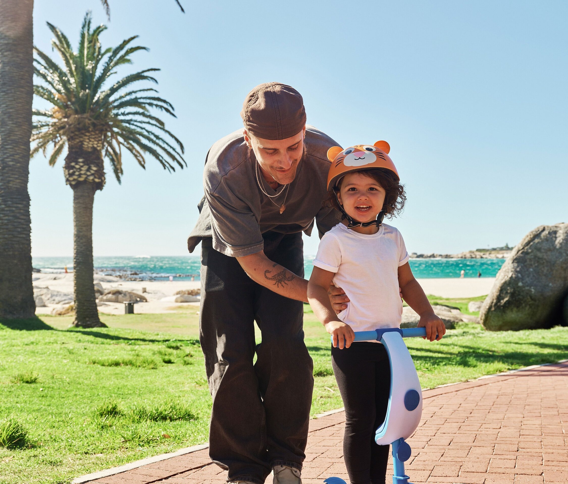 Mann hilft einem Kind mit Tigerhelm auf einem blauen Hudora 4-in-1-Scooter. Palmen und Meer im Hintergrund.