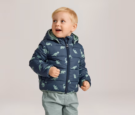 Kleiner Junge mit blonden Haaren trägt eine marineblaue, gesteppte Jacke mit Dinosaurier-Muster und grüne Hosen.