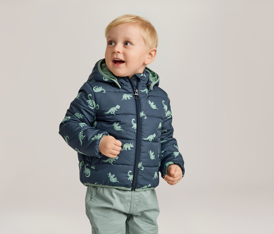 Kleiner Junge mit blonden Haaren trägt eine marineblaue, gesteppte Jacke mit Dinosaurier-Muster und grüne Hosen.