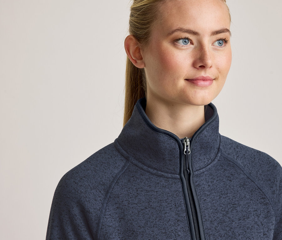 Nahaufnahme einer Frau mit blonden Haaren und blauen Augen, die eine dunkelblaue Fleecejacke mit Reißverschluss und Stehkragen trägt.
