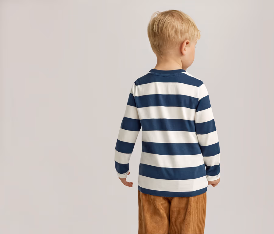 Rückansicht eines Kindes mit blonden Haaren in einem blau-weiß gestreiften Langarmshirt und brauner Cordhose vor einem hellen Hintergrund.
