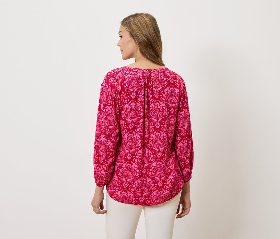 Rückansicht einer Frau mit blonden Haaren, die eine pinkfarbene, langärmlige Bluse mit Muster und weiße Hosen trägt.