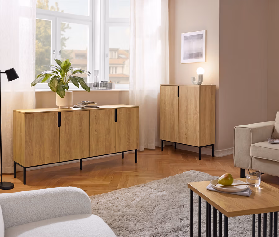 Ein Wohnzimmer mit einem langen Sideboard aus hellem Holz unter einem Fenster und einem passenden hohen Schrank. Eine schwarze Stehlampe, zwei Sessel, ein zotteliger Teppich und ein Couchtisch mit einer Birne und einem Wasserglas sind ebenfalls zu sehen.