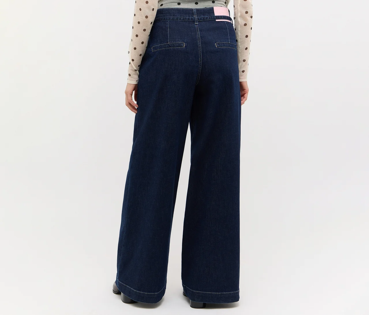 Person von hinten in dunkelblauer weiter Jeans und einem hellen, gepunkteten Oberteil. Ein pinkes Etikett ist am hinteren Bund der Jeans sichtbar.