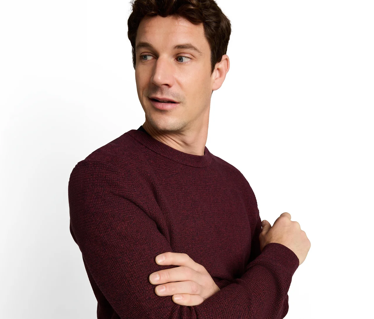 Mann mit braunen Haaren und blauen Augen, der einen bordeauxroten, strukturierten Pullover trägt, blickt mit leicht geöffnetem Mund nach rechts. Seine Arme sind vor ihm verschränkt.