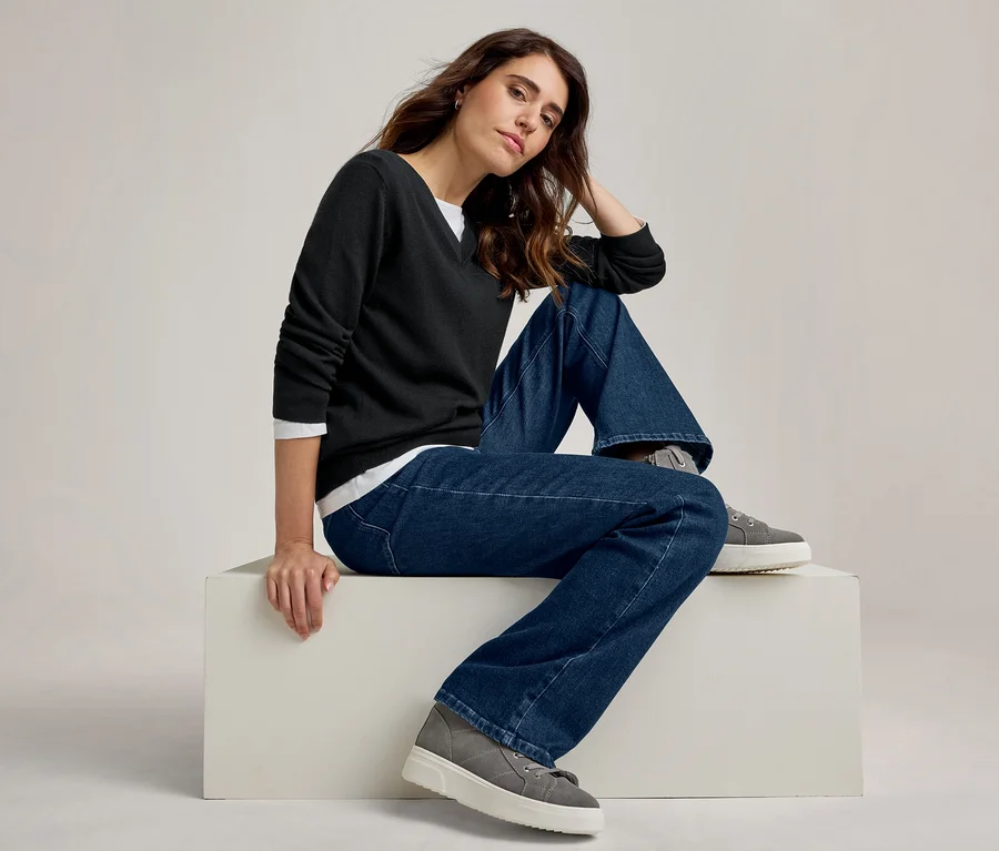Frau sitzt auf einem weißen Block, trägt einen schwarzen V-Pullover über einem weißen Shirt, weite blaue Jeans und graue Plateau-Sneaker.