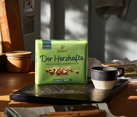 Auf einem Tisch steht eine Packung Der Herzhafte - 500 g Gemahlen Kaffee neben einer Tasse und einem Sandwich.