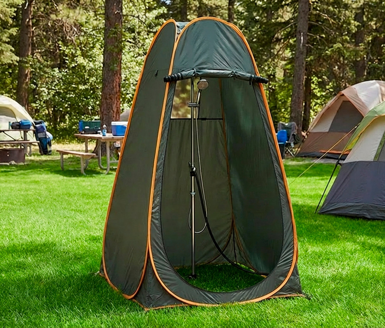 Ein mobiles Dusch- und Umkleidezelt in Dunkelgrün mit orangefarbenen Akzenten steht auf grünem Gras auf einem Campingplatz. Im Zelt ist ein Duschkopf sichtbar. Im Hintergrund sind weitere Zelte und Picknicktische unter Bäumen zu sehen.
