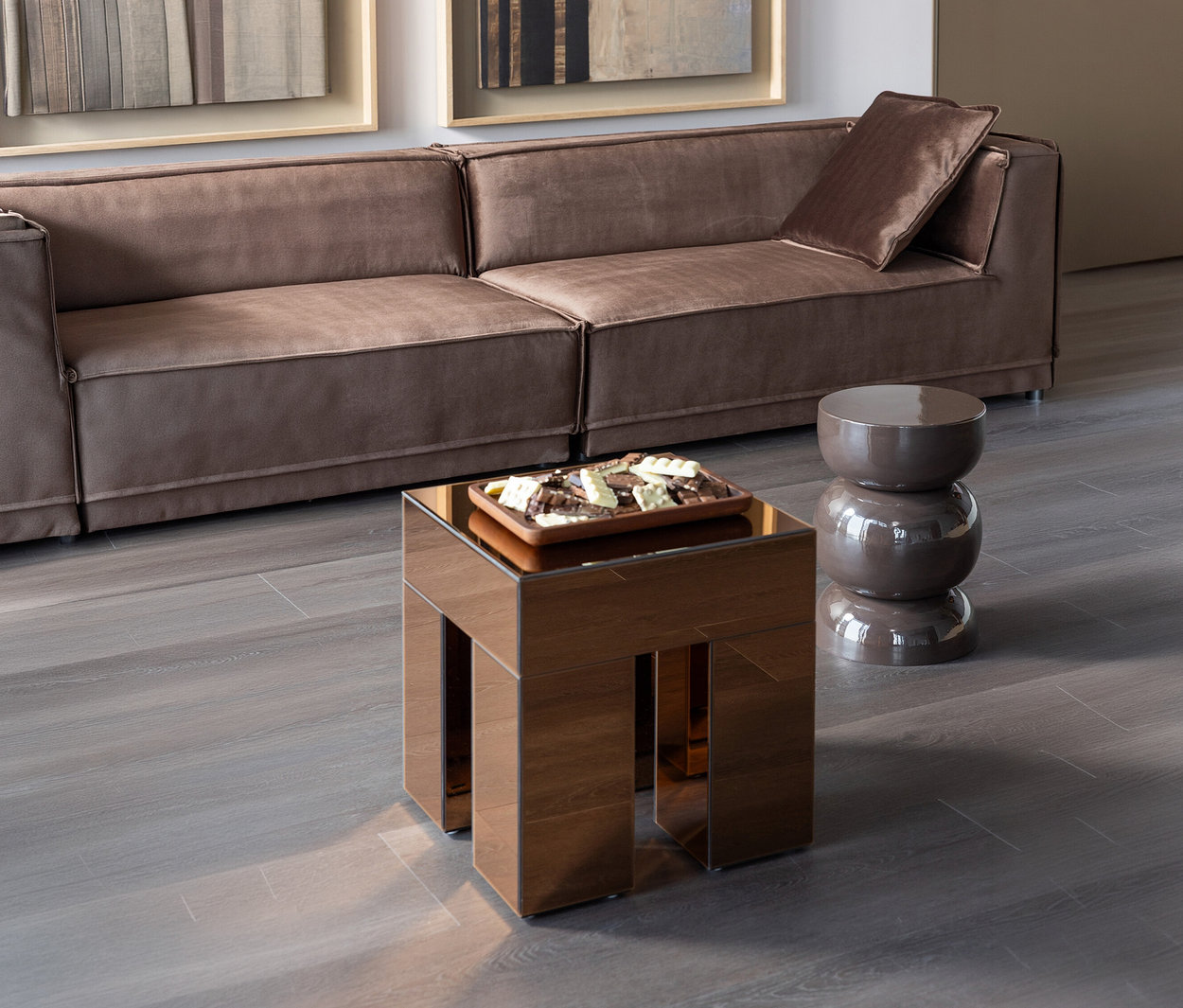 Ein modernes Wohnzimmer mit einem langen, braunen Modulsofa, einem quadratischen Spiegeltisch mit einer Schale Pralinen und einem glänzenden, dunkelbraunen Hocker in Sanduhrform auf einem grauen Holzboden.