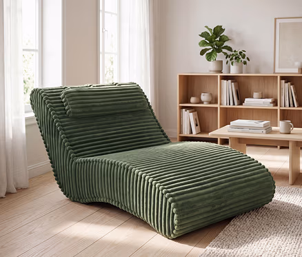 Grüne Cord-Chaiselongue mit gerippter Textur und Kopfstütze in einem hellen Wohnzimmer mit Holzregal, Büchern und Fenster.
