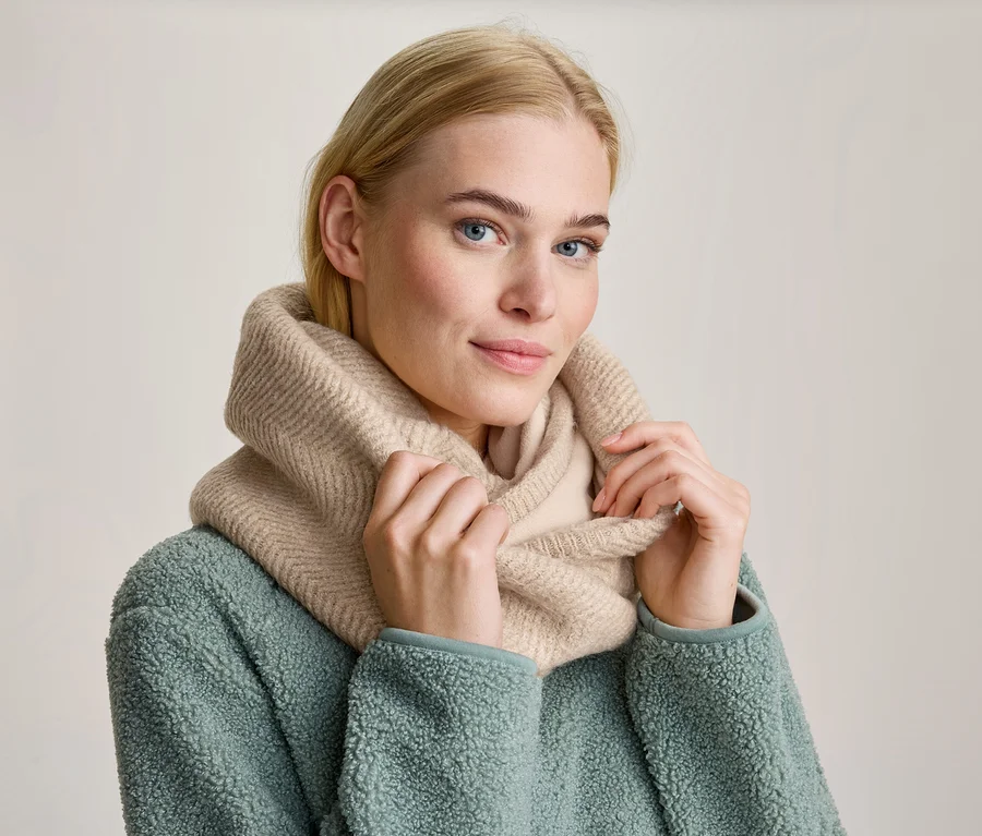 Blonde Frau mit blauen Augen trägt einen beigefarbenen Strick-Rundschal und eine mintgrüne Fleecejacke, hält den Schal mit beiden Händen und blickt nach vorne.