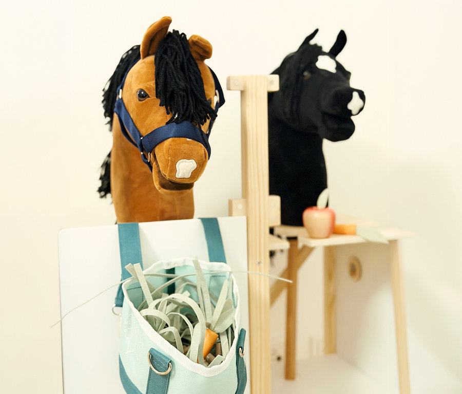 Ein small foot Hobby-Horse-Pferdestall mit einem braunen und einem schwarzen Steckenpferd. Ein Sack mit Spielzeug-Heu hängt am Stall.