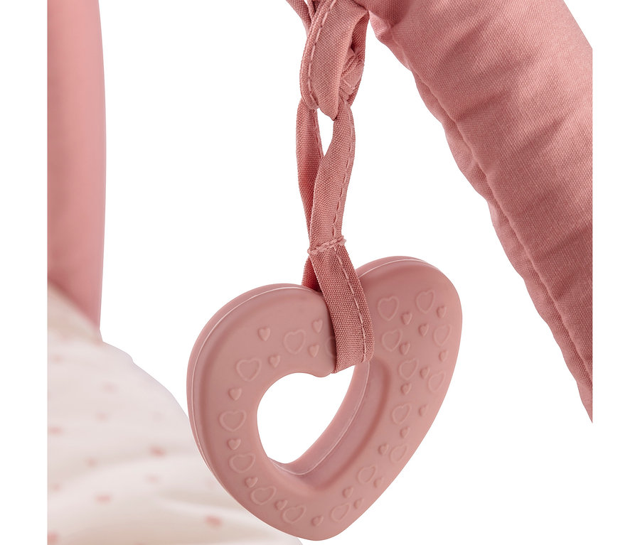 Ein rosafarbener herzförmiger Beißring mit kleinen geprägten Herzen hängt an einem rosafarbenen Stoffband, befestigt an einer gepolsterten rosa Stange. Im Hintergrund ist ein weißer Stoff mit rosa Punkten zu sehen.