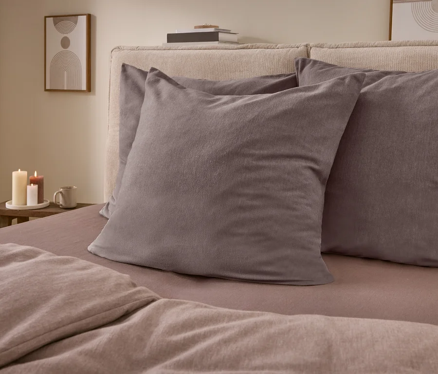Ein Bett mit hell gepolstertem Kopfteil, zwei dunkelgrauen Kissen und hellbrauner Bettwäsche. Daneben ein Holztisch mit drei brennenden Kerzen, einer Tasse und einem gerahmten Bild an der Wand.