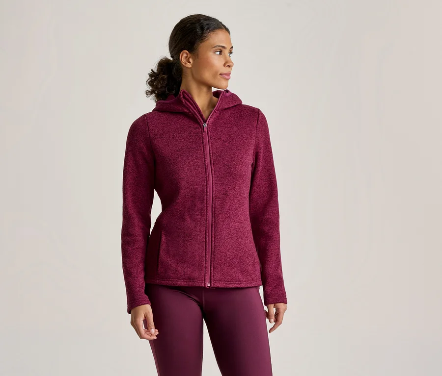Eine Frau trägt eine burgunderfarbene Kapuzenjacke mit durchgehendem Reißverschluss und passende Leggings, steht und blickt nach rechts.